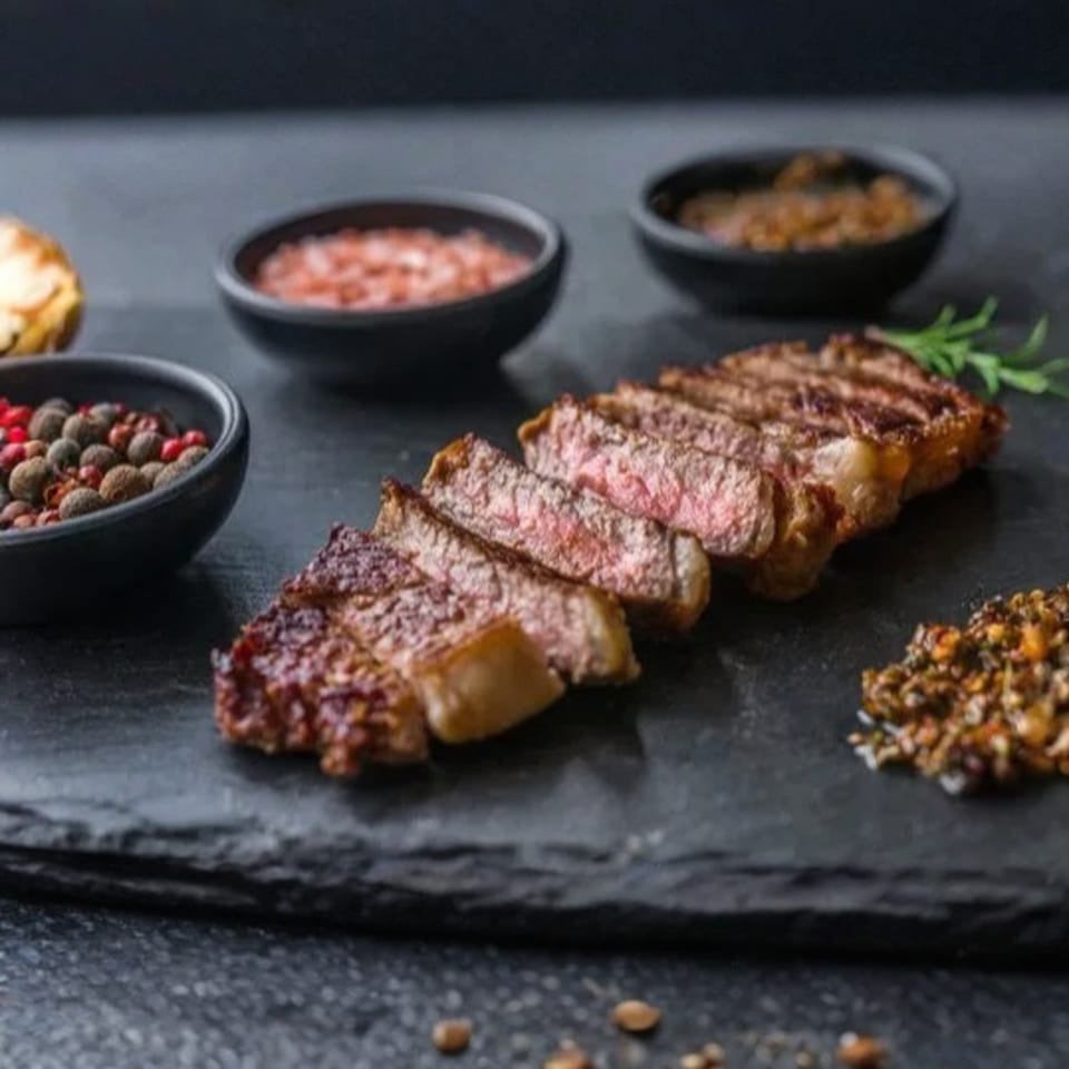 Sliced steak with various spices and sauces on a dark stone board.