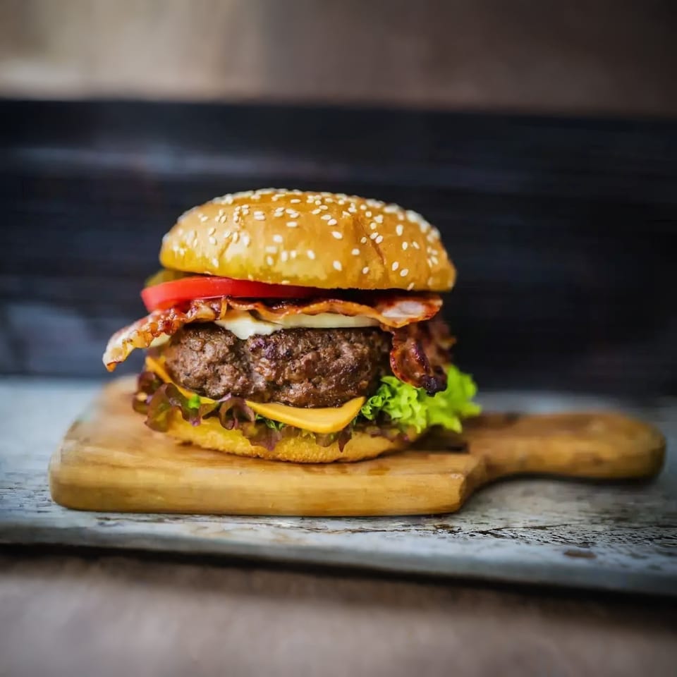 A juicy bacon cheeseburger with lettuce and tomato on a sesame seed bun, served on a wooden board.