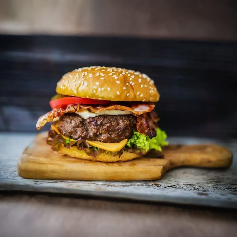 A juicy bacon cheeseburger with lettuce and tomato on a sesame seed bun, served on a wooden board.